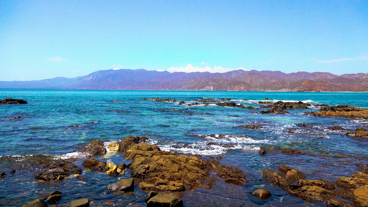 Small waves break around foreground rocks beneath a blue sky.