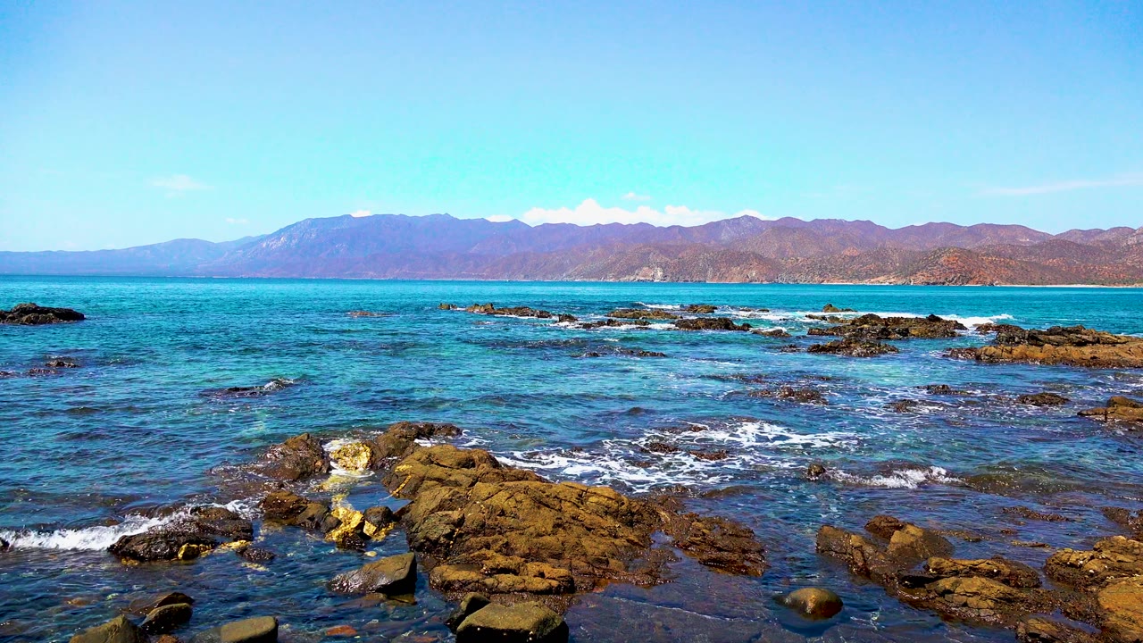 A bright El Sargento La Ventana shoreline view with blue-green water crossing low reef rocks, brown landforms on the horizon, and no visible people.