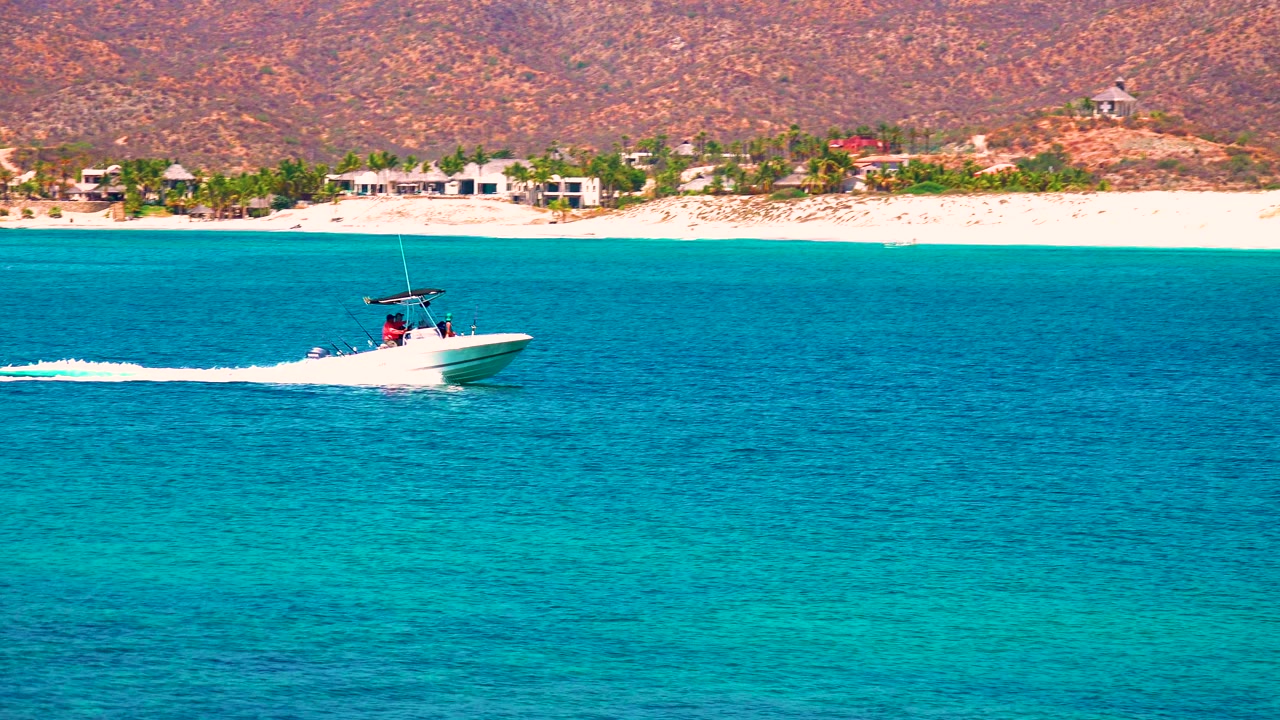 At El Sargento La Ventana, Mexico, the boat sits farther across the frame on deep blue water, with its wake trailing left and a pale beach and hillside settlement in the distance.