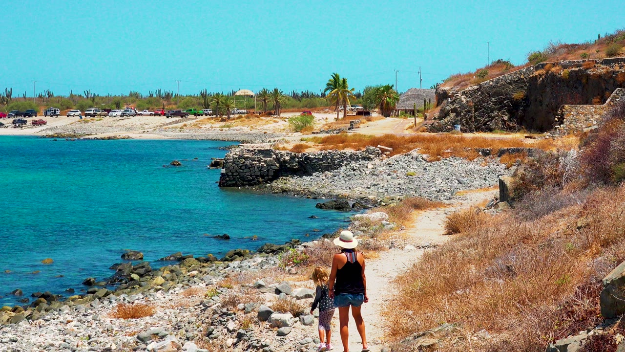 In El Sargento La Ventana, Mexico, a woman in a sun hat stands on a pale rocky path while a child and small brown dog face the turquoise water at the start of the clip.