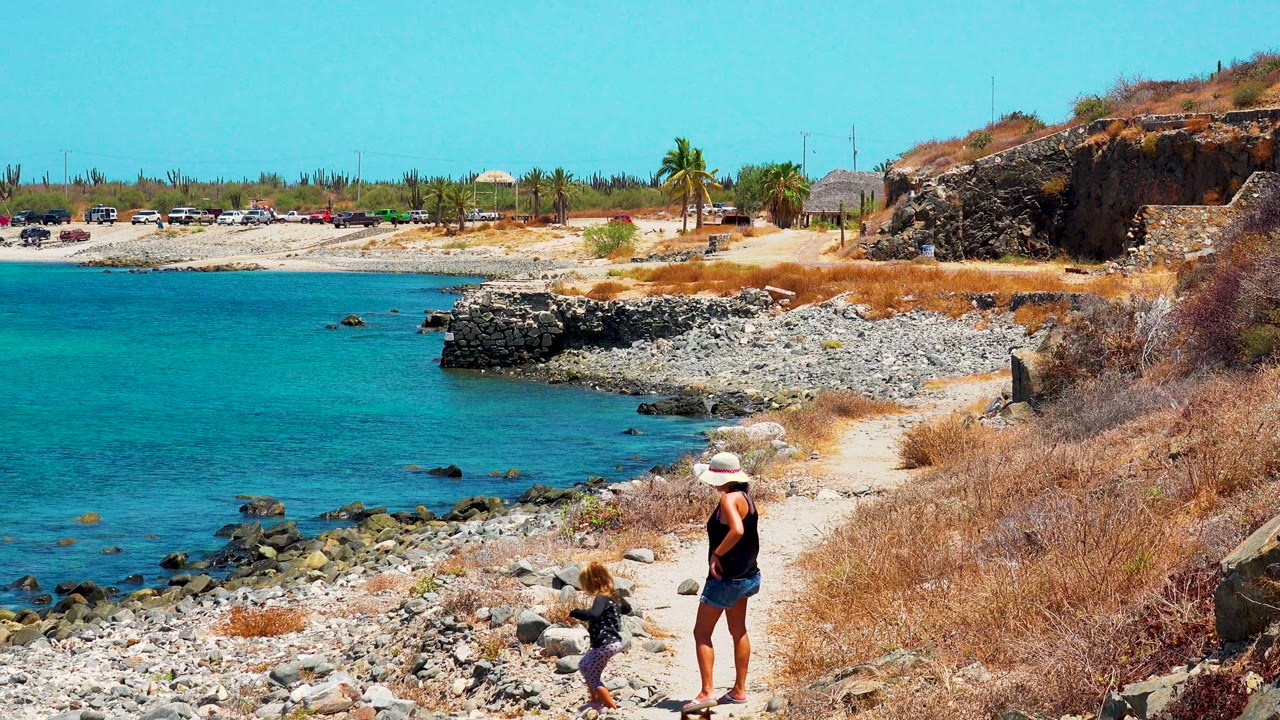 A child in dark clothing steps near the woman on the rocky shoreline path while the dog moves nearby and the blue water, stone point, and dry hillside sit behind them in Mexico.