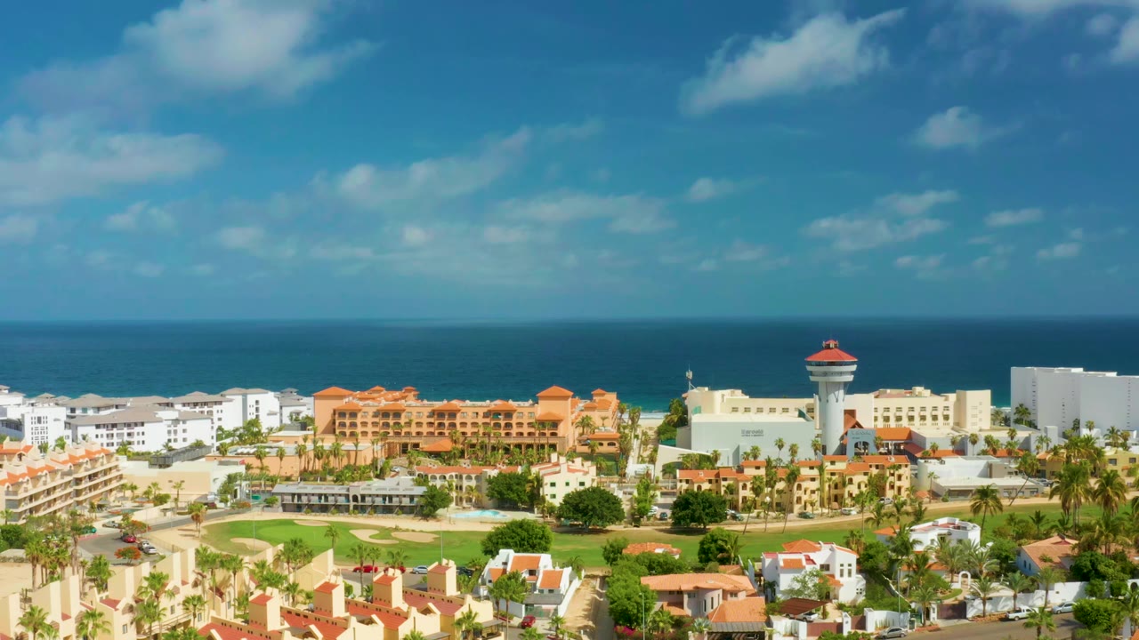San Jose Del Cabo, Mexico seen from above with terracotta-roofed buildings centered against the ocean and a bright clouded sky, no visible people.