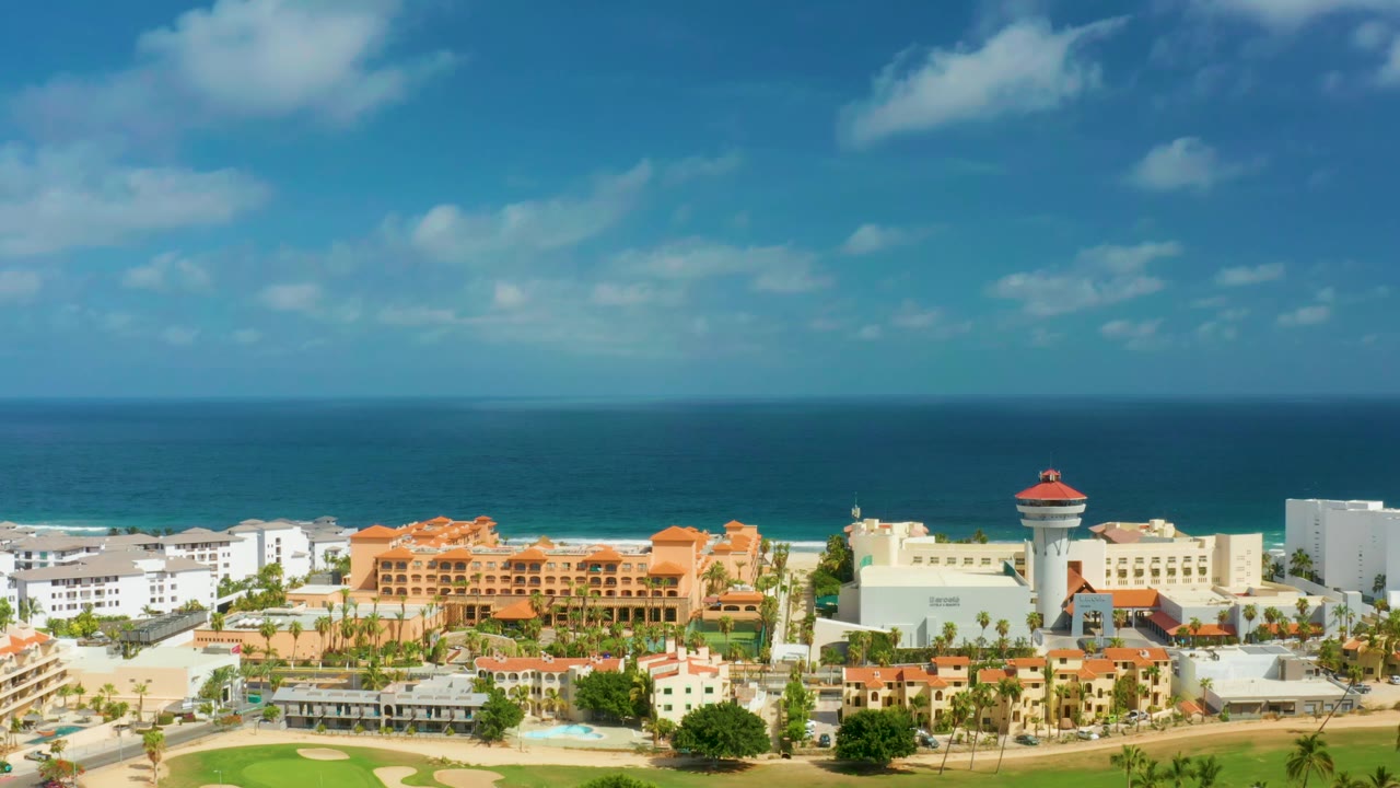 Aerial view in San Jose Del Cabo with the resort district spread across the lower frame, surf behind the buildings, a broad blue ocean band, cloud patches, and no visible people.