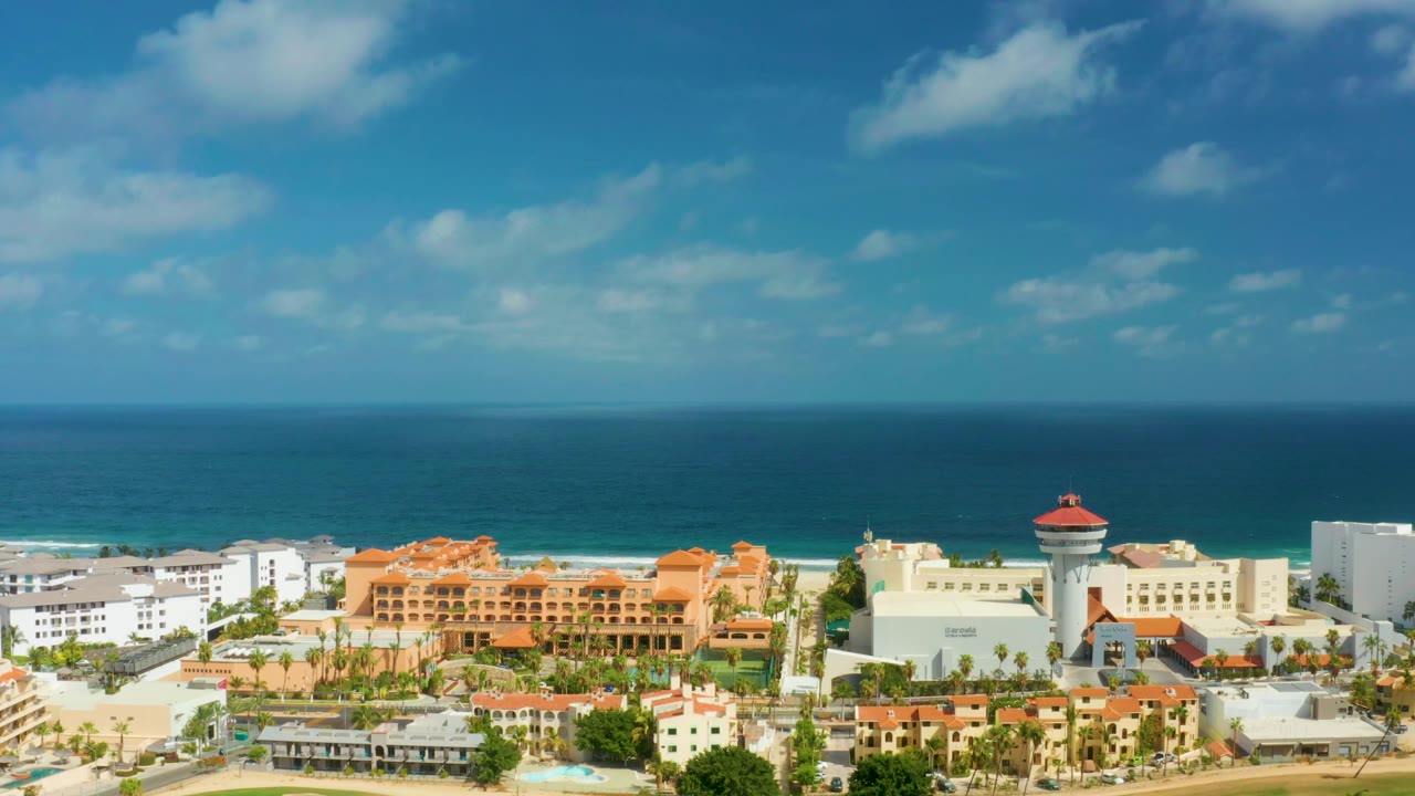 San Jose Del Cabo resort buildings sit below a bright blue sky, with a sandy beach and white surf separating the hotels from the darker offshore water and no visible people.