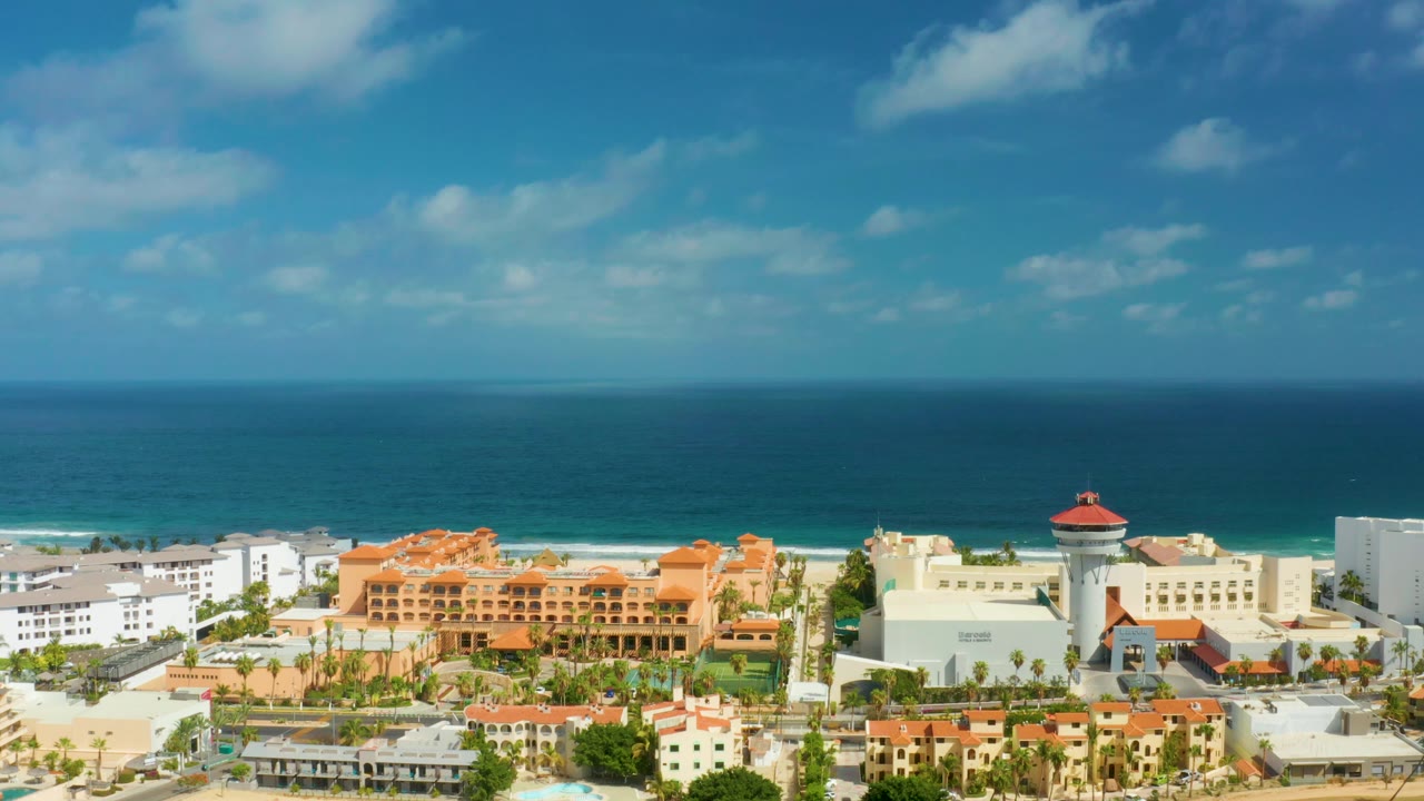A high aerial frame in San Jose Del Cabo shows large resort buildings, palms and landscaped grounds, a narrow sandy beach, turquoise shoreline water, open ocean, and no visible people.