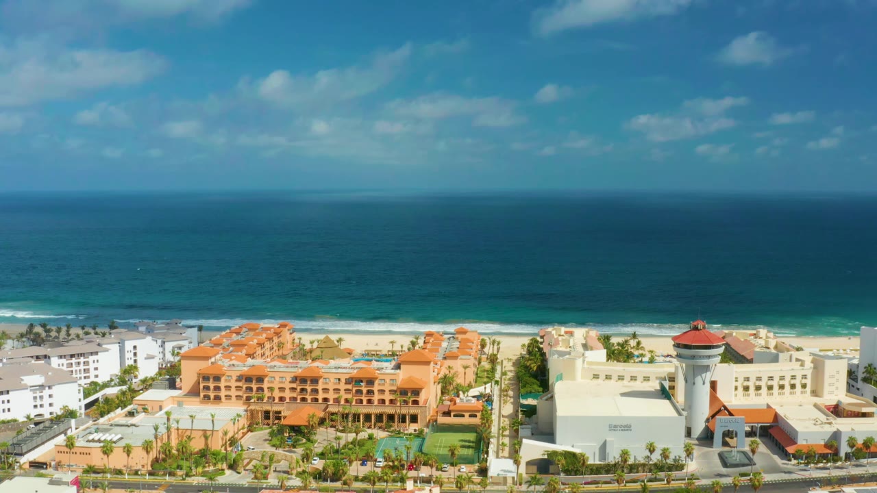 Wide elevated view in San Jose Del Cabo, Mexico with the ocean horizon high in frame, surf lines along the sand, resort pools, orange roofs, and no visible people.