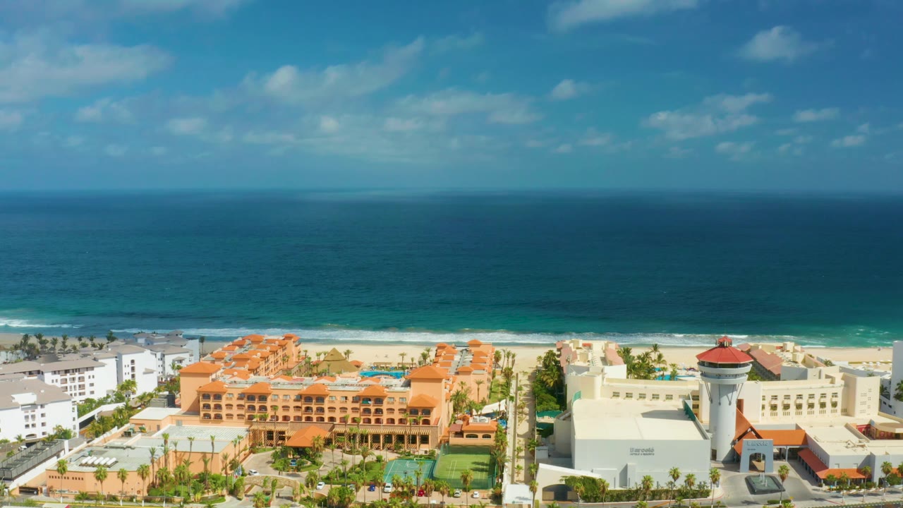 Aerial frame in San Jose Del Cabo, Mexico showing a beach resort complex with orange roofs, a red-roofed tower, white hotel structures, blue ocean, small clouds, and no visible people.