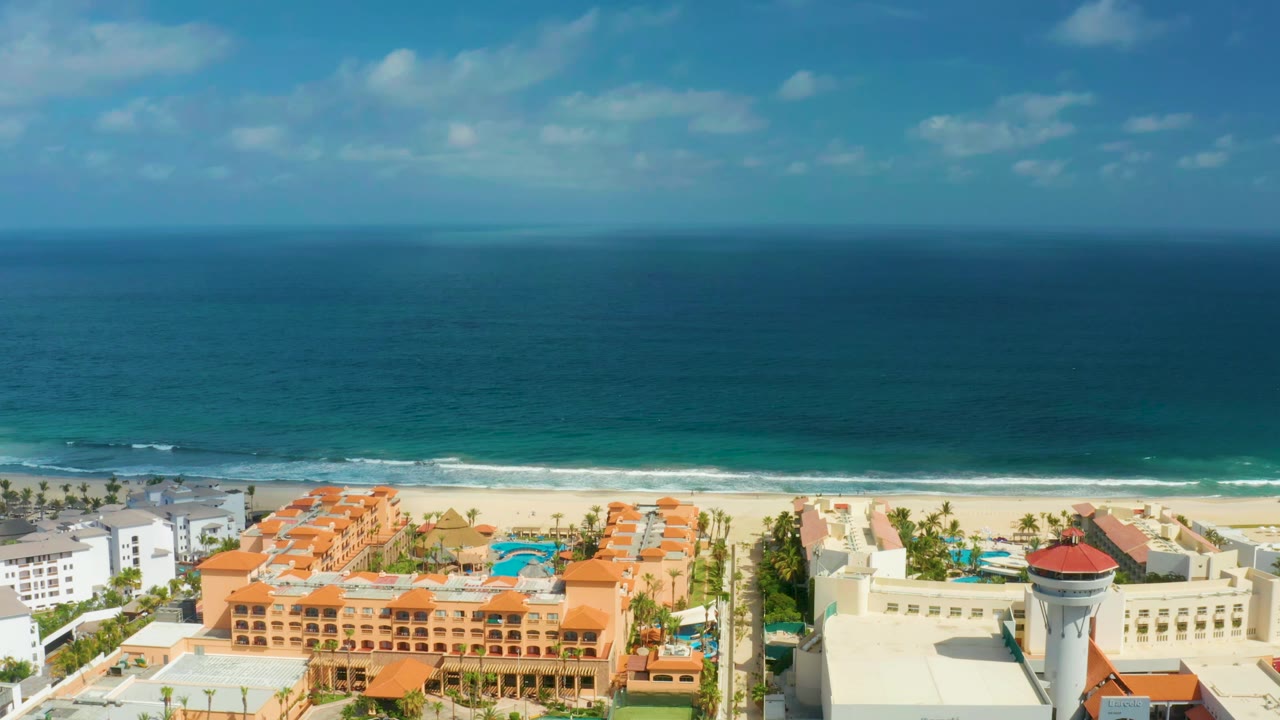 San Jose del Cabo coastline frame with orange resort roofs and turquoise pool areas in the lower frame, pale sand and white waves across the middle, and no visible people.