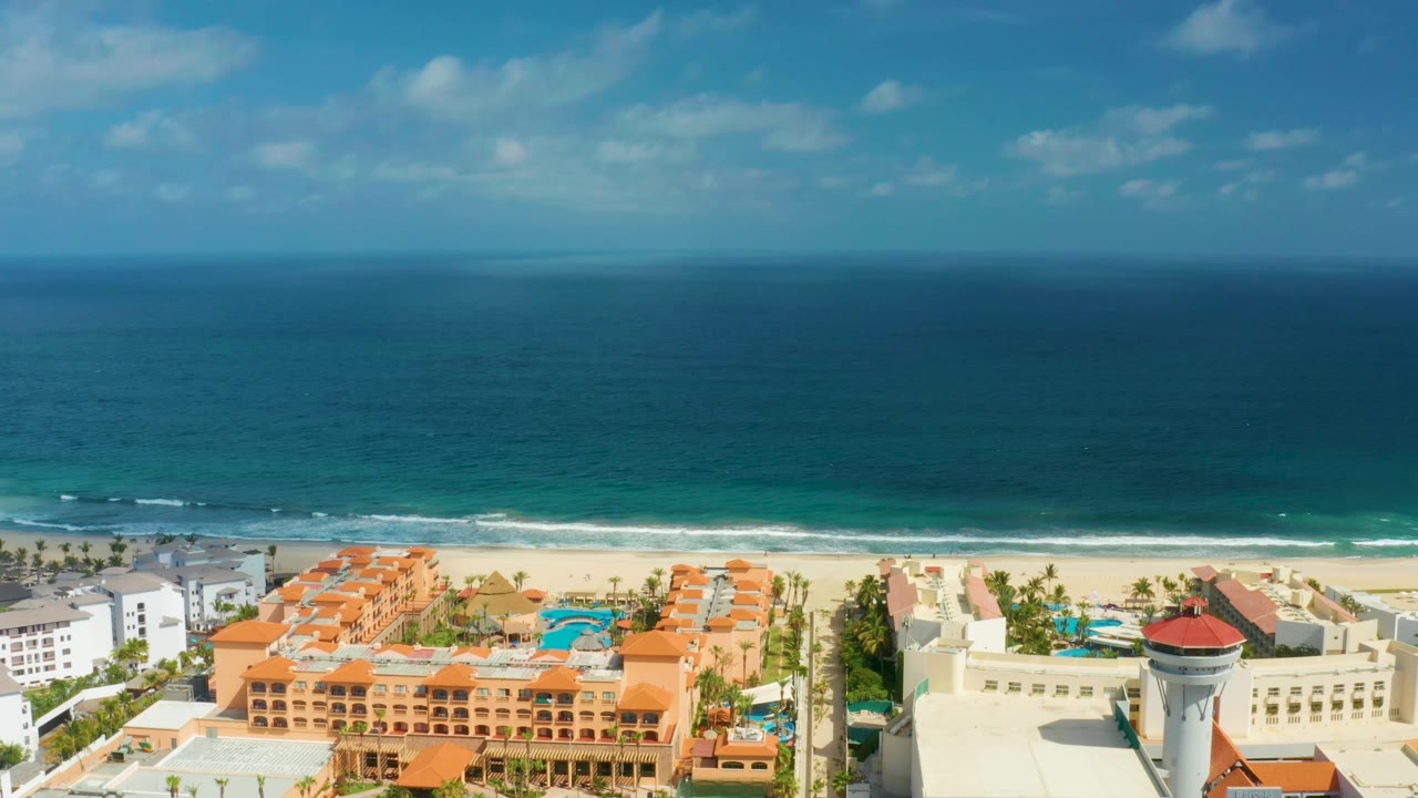 Elevated San Jose del Cabo view where a cream building and red-roofed tower sit near orange resort blocks, with the blue sea and low surf behind them and no visible people.