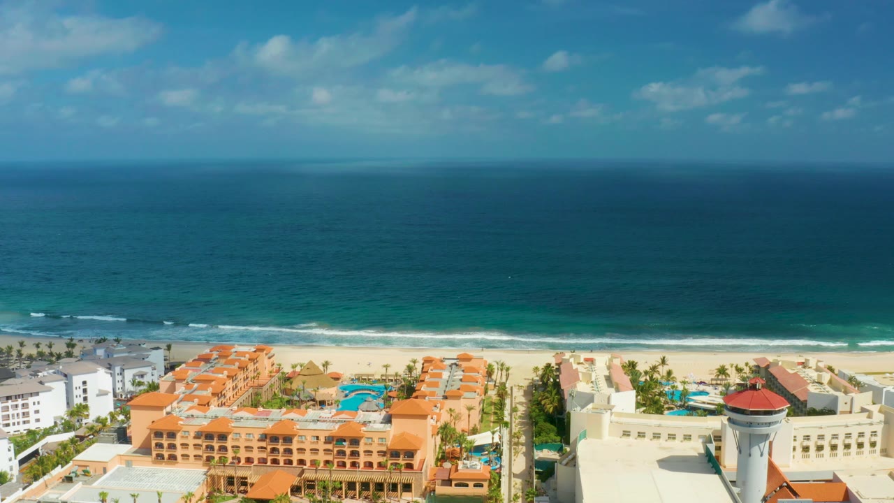Mid-clip frame over San Jose del Cabo resorts, with the horizon high in the image, small wave breaks near the beach, orange roofs at the bottom edge, and no visible people.