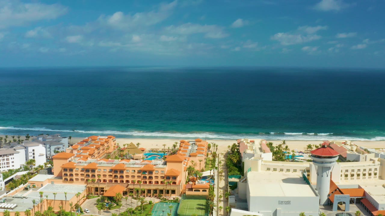 Aerial frame over San Jose Del Cabo resort buildings with orange roofs, sandy beach, blue ocean horizon, and no visible people.