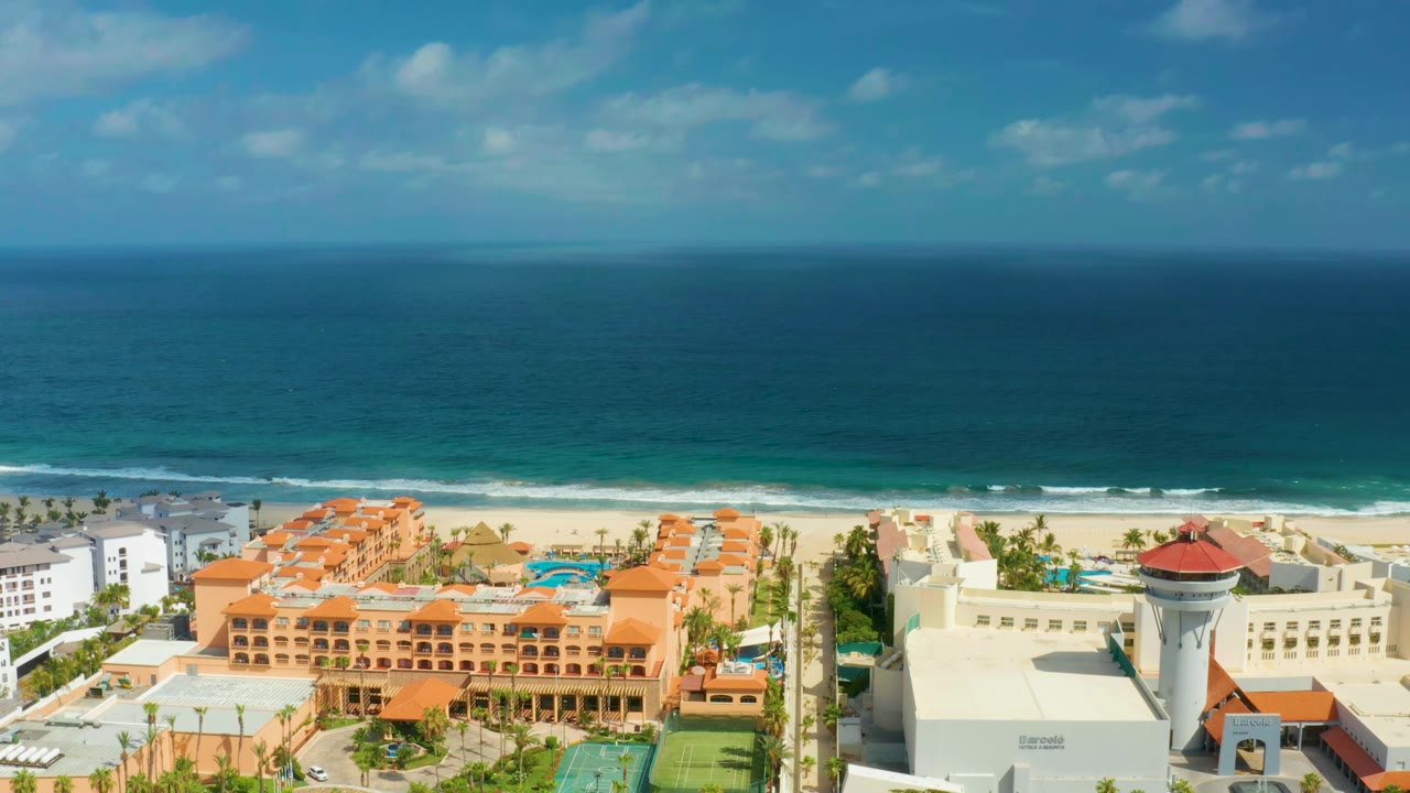 High aerial view in San Jose Del Cabo showing orange beachfront resort blocks, a pale beach strip, rolling surf, and no visible people.