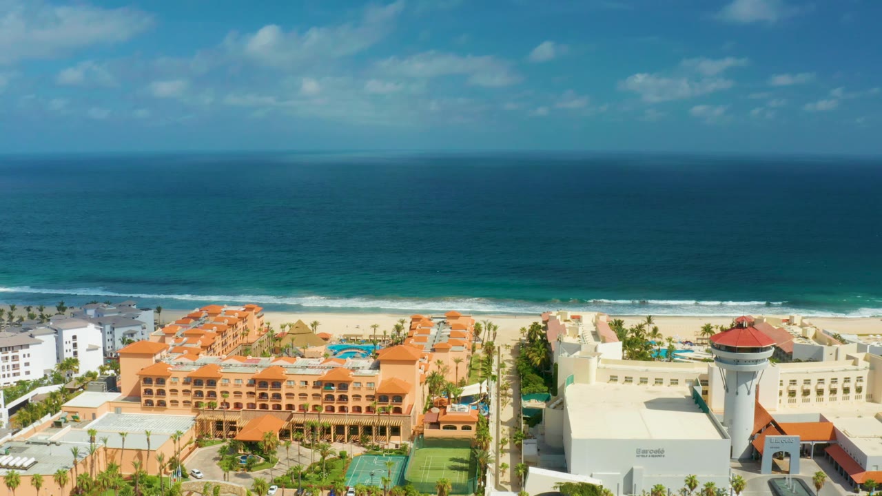Aerial San Jose Del Cabo frame with a white tower-like resort structure, orange hotel buildings, beach, ocean horizon, and no visible people.