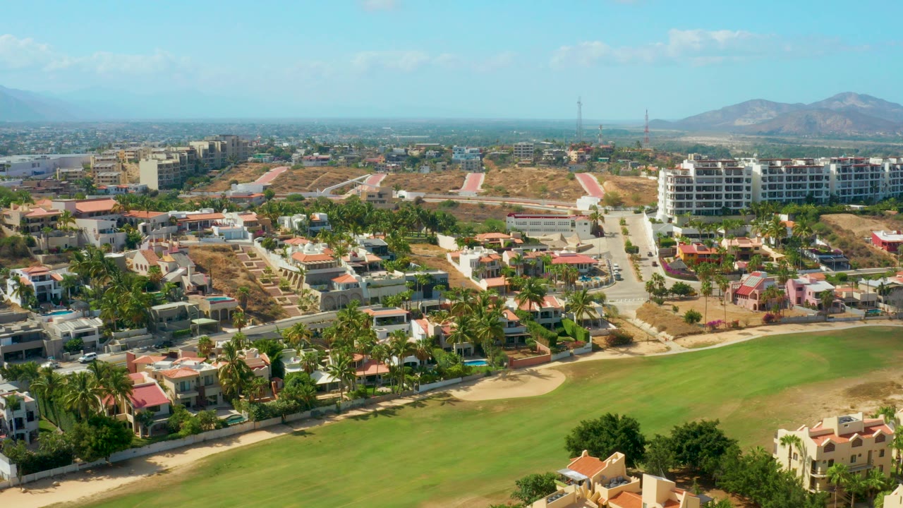 San Jose Del Cabo aerial view with a green fairway in the foreground, residential roofs and palm trees across the middle, hills and mountains in the distance, and no visible people.