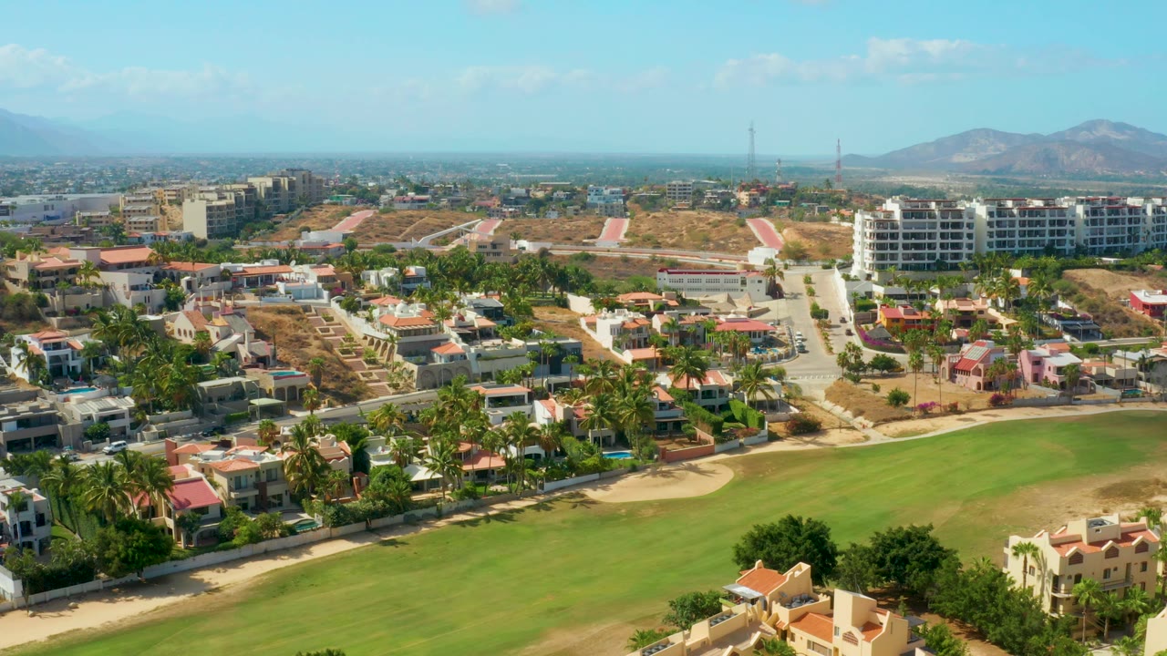 San Jose Del Cabo aerial frame showing low homes, a curved fairway edge, sparse dry ground, modern buildings on the hill, and distant mountains under clear daylight, with no visible people.