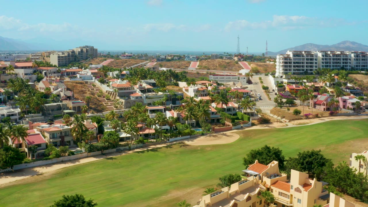 Opening aerial frame in San Jose del Cabo, Mexico, looking across a green field toward residential homes, palms, distant buildings, low hills, and no visible people.