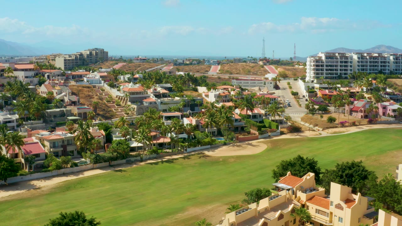 Aerial view in San Jose del Cabo with the grassy field filling the lower frame, palm-lined homes across the center, larger white buildings near the horizon, and no visible people.