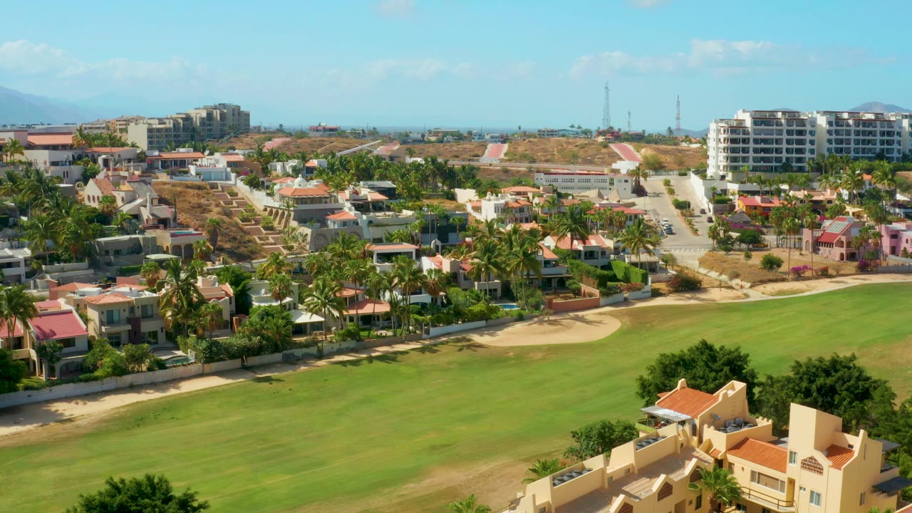 Wide aerial view of field, houses, palms, roads, and low hills under blue sky.