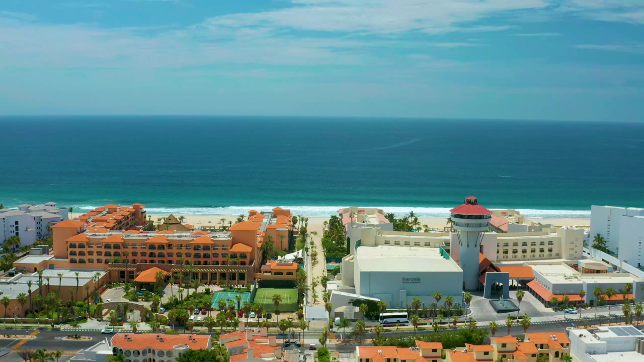 Aerial frame in San Jose Del Cabo, Mexico with the ocean horizon high in view, surf line along the beach, resort blocks, roadside vehicles, palms, and no visible people.