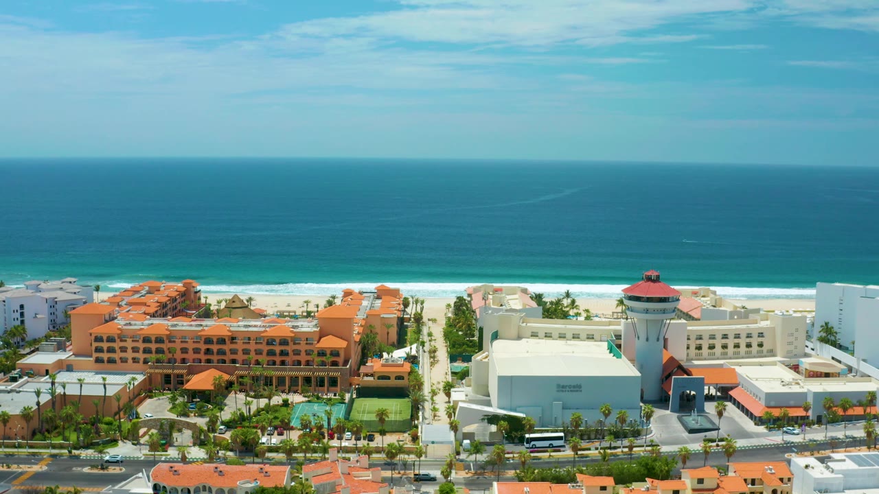 Wide aerial frame in San Jose Del Cabo, Mexico showing turquoise ocean bands, a bright beach strip, orange resort roofs, a round-topped tower-like building, roads, and no visible people.