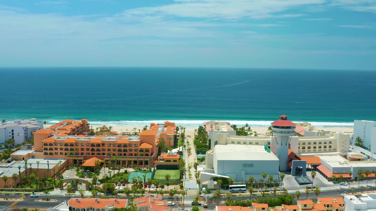 San Jose Del Cabo aerial view with broad ocean, white surf, beachfront hotels, orange rooftops, a pale modern building, a road at the bottom edge, and no visible people.