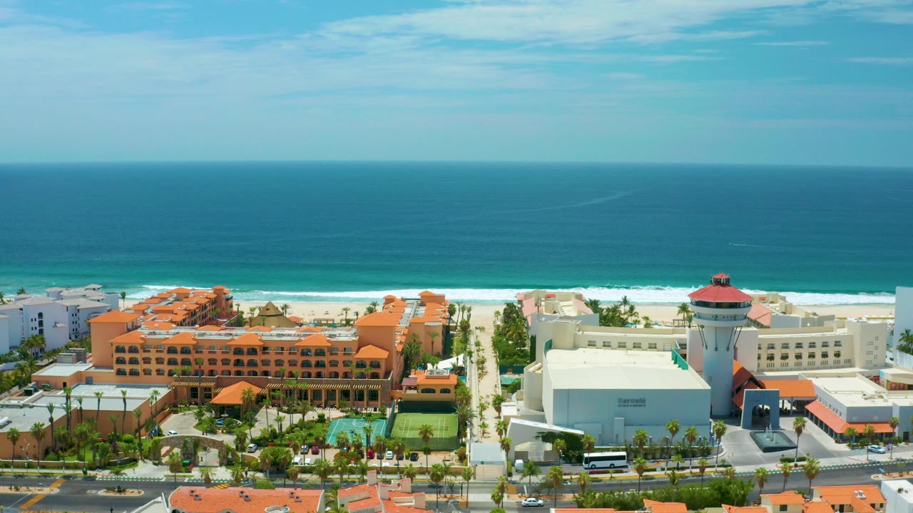 Aerial view in San Jose Del Cabo, Mexico showing orange-roofed resort buildings, tennis courts, a white tower, sandy beach, blue ocean, and no visible people.