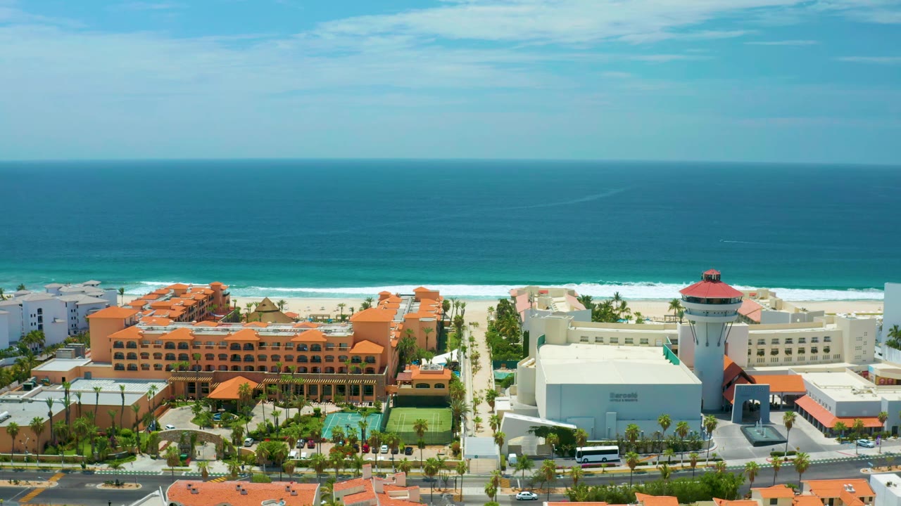 High coastal view in San Jose Del Cabo, Mexico with orange hotel roofs, a visible pool courtyard, pale beach, ocean bands, and no visible people.