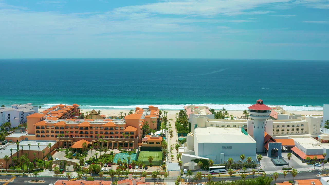 Aerial resort view in San Jose Del Cabo, Mexico with the white red-roofed tower more prominent beside orange-roofed hotel buildings, beach, ocean, and no visible people.