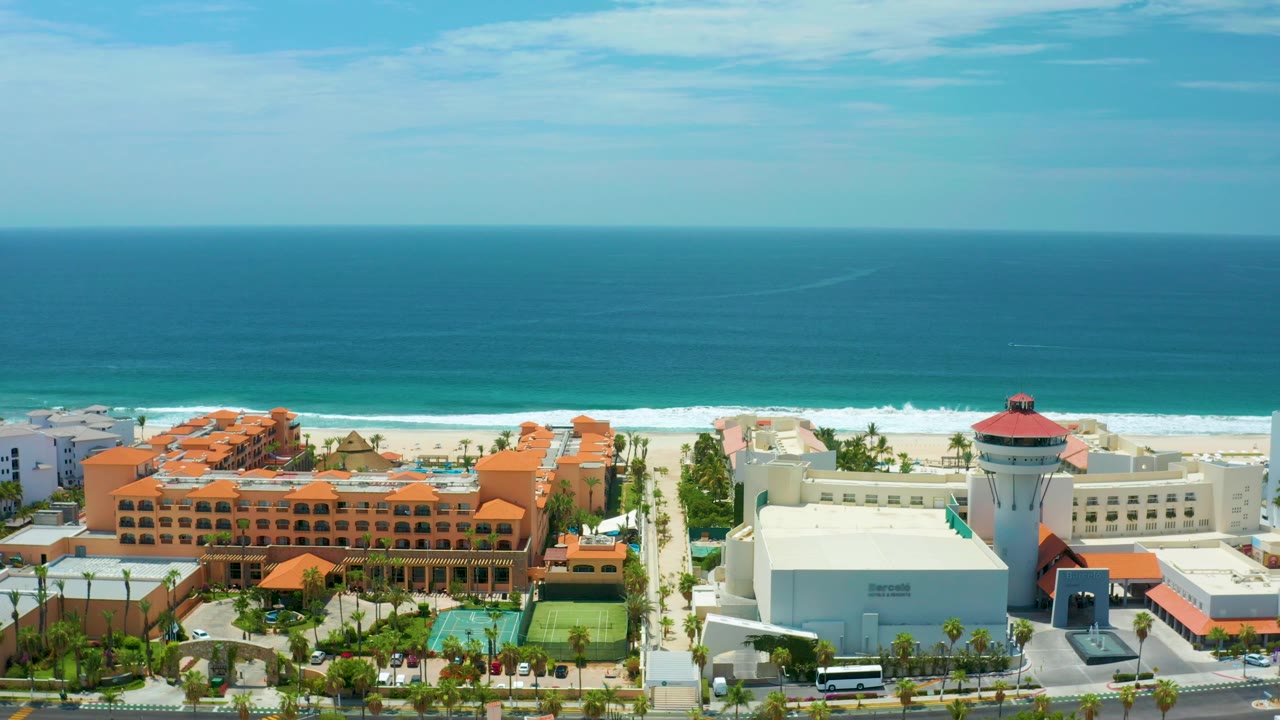 San Jose Del Cabo aerial frame showing the resort frontage shifted across the composition, with tennis courts, palms, sand, blue water, and no visible people.