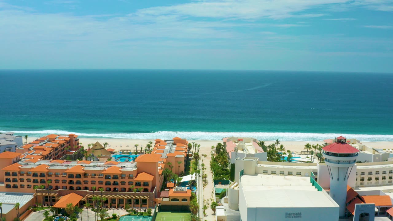 High aerial view over San Jose Del Cabo resort roofs, palm rows, pools, white beach surf, and turquoise ocean under bright sky, with no visible people.