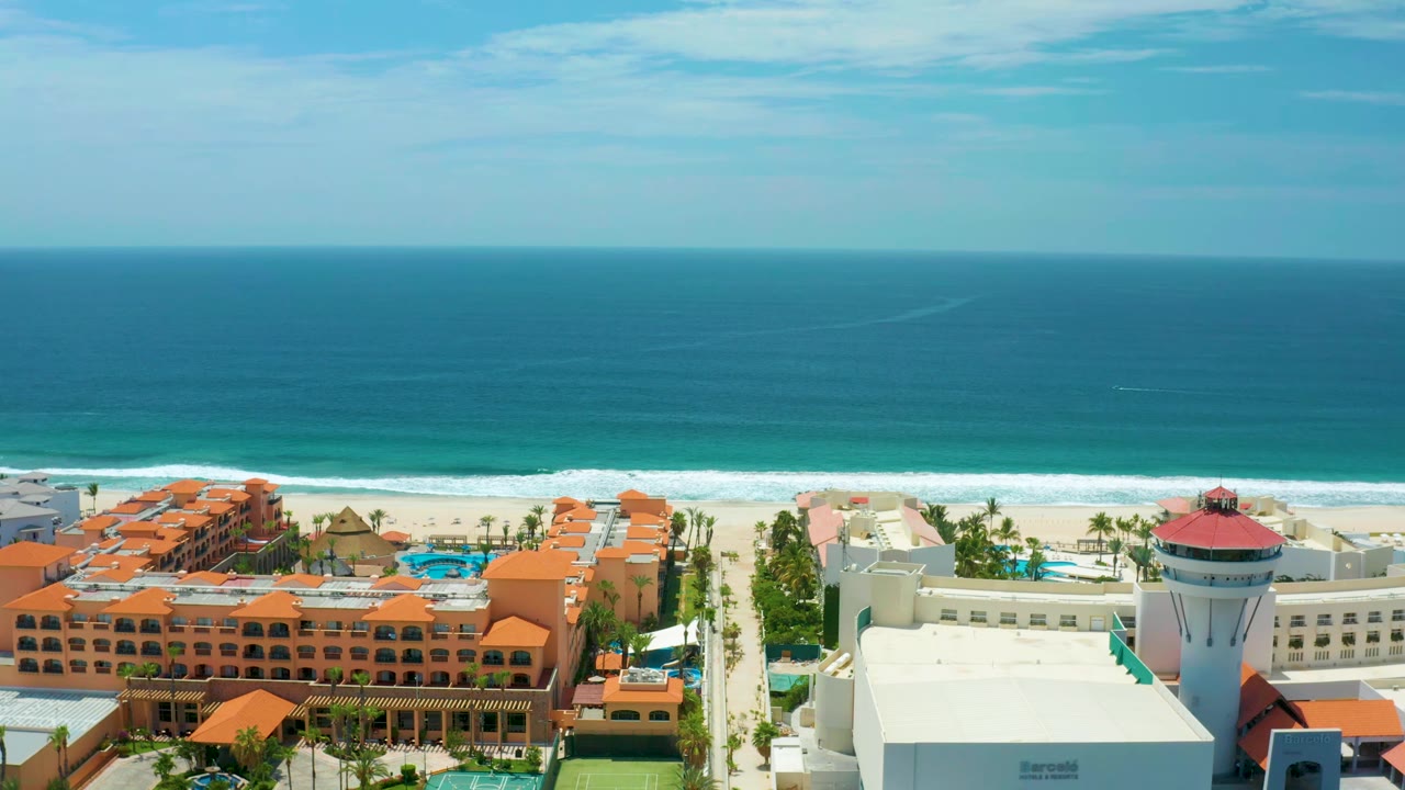 Aerial frame in San Jose Del Cabo showing orange hotel roofs, blue pools, palms, pale beach sand, and a long band of breaking surf, with no visible people.