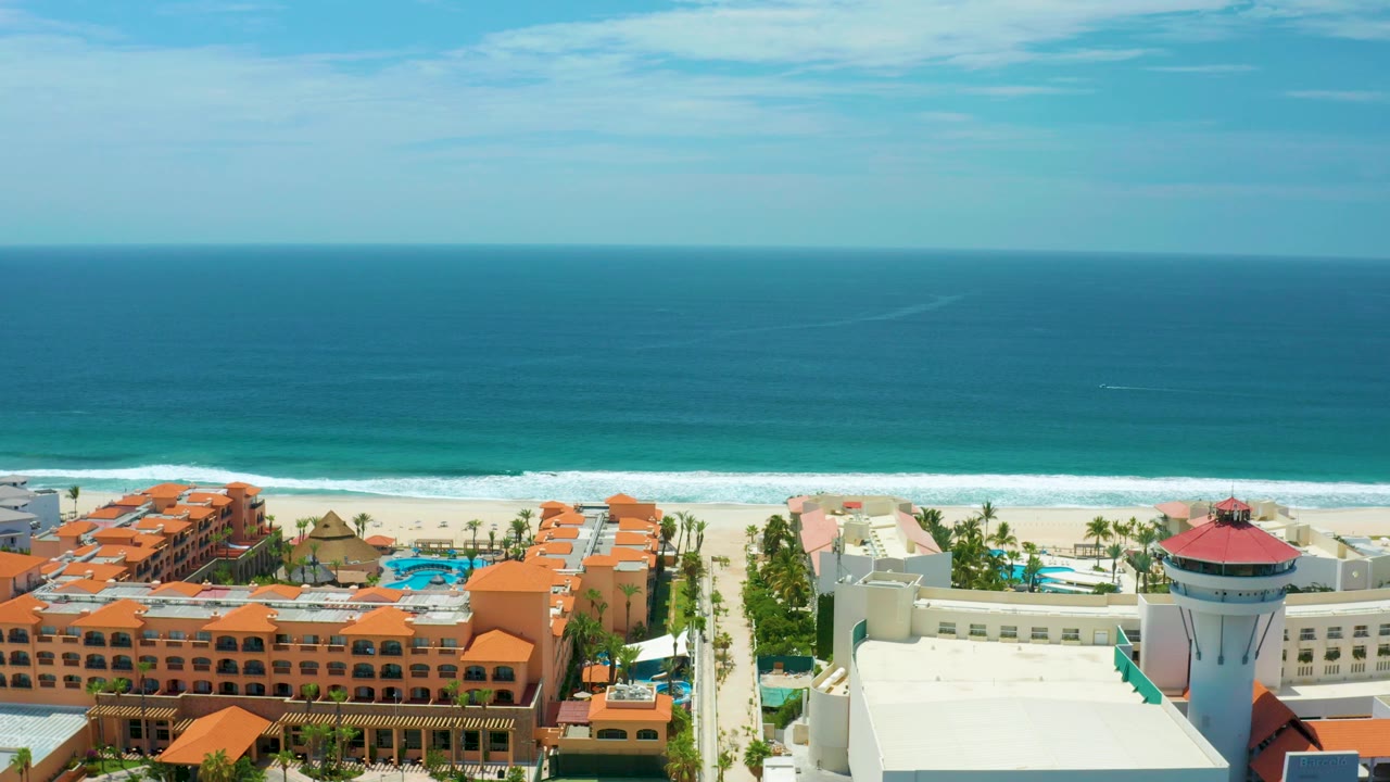 San Jose Del Cabo aerial view with a white resort block and round red-roof tower in the foreground, orange resort buildings to the left, surf beyond, and no visible people.