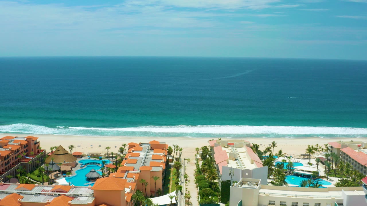 Aerial view over San Jose Del Cabo beachfront residences with orange roofs, pools, palms, pale sand, white surf, blue ocean, and no visible people.