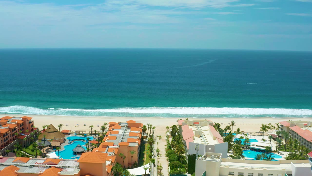 Wide aerial coastline with orange-roofed residences, palms, pools, beach, and ocean.