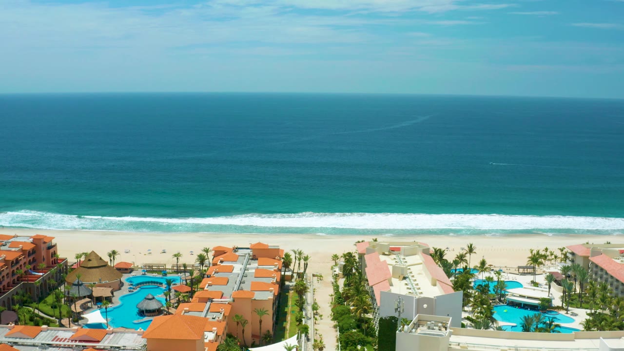 Aerial view in San Jose Del Cabo with cream and orange beachfront buildings, turquoise swimming pools, palm rows, white surf, open ocean, and no visible people.