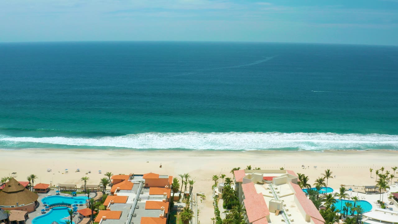 Opening aerial frame over a San Jose Del Cabo beach with turquoise water, a white surf line, pale sand, resort pools and orange roofs along the lower edge, and no visible people.