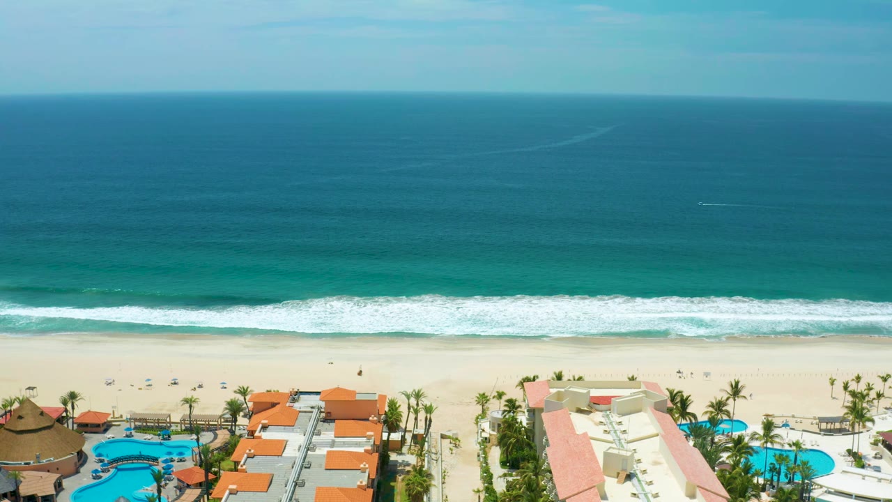 Wide aerial coastline with waves breaking toward a resort beach and pool area.