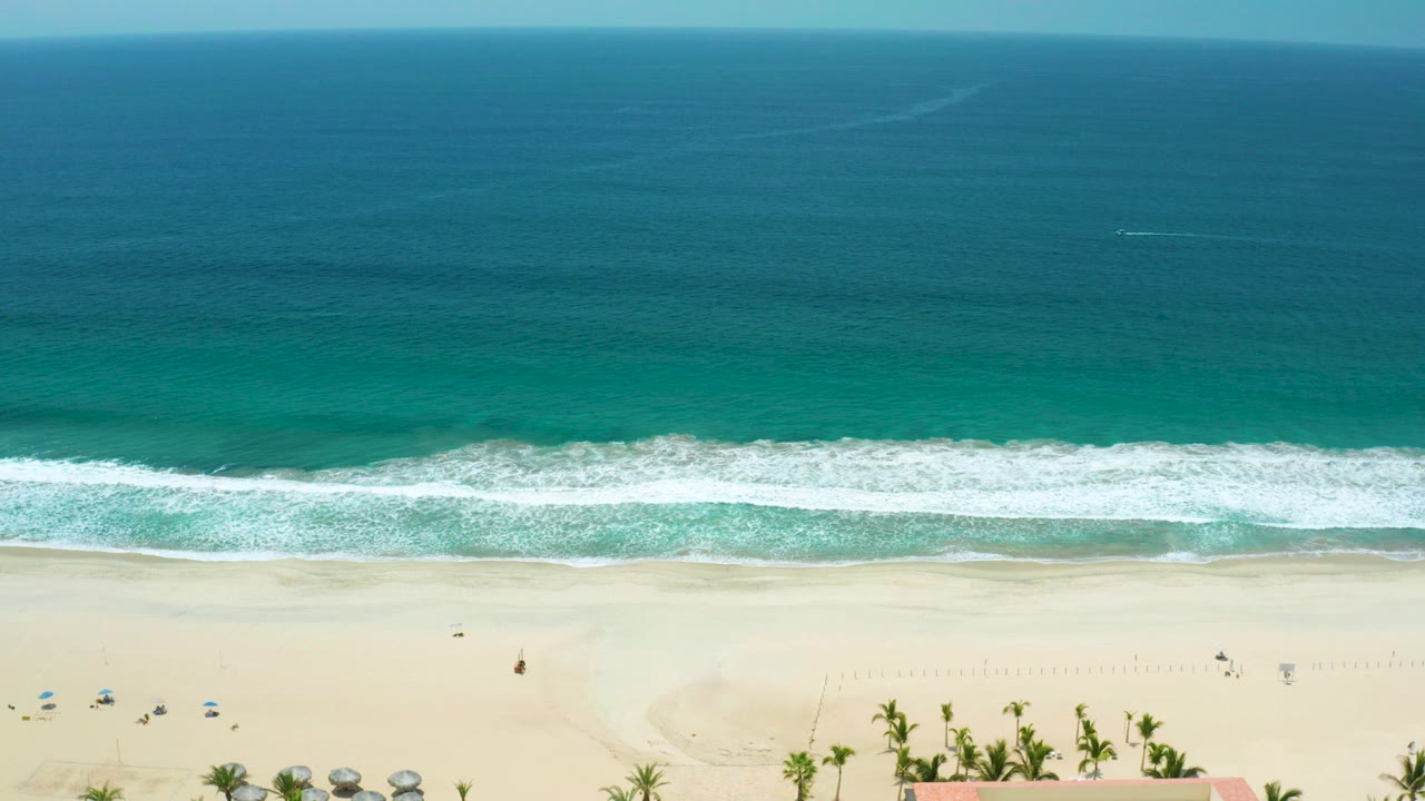 Wide aerial frame over San Jose del Cabo, Mexico, showing teal ocean, a white surf line, pale sand, small umbrellas near the lower left edge, and no visible people.