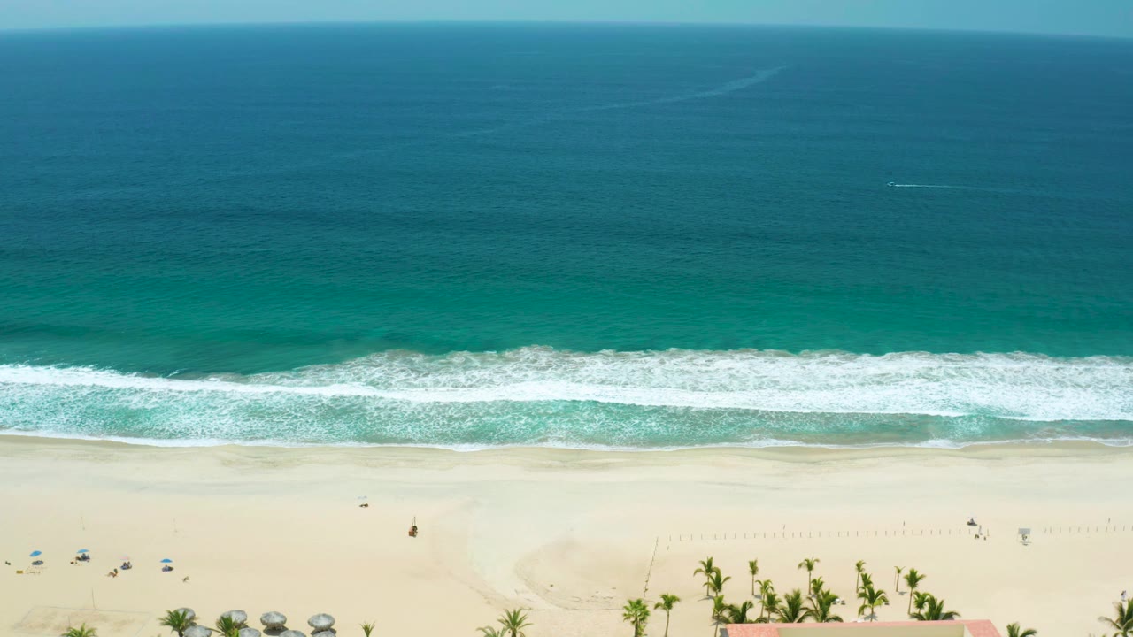Aerial view in San Jose del Cabo, Mexico, with the white wave band shifted across the center, broad teal water above it, pale sand below, and no visible people.