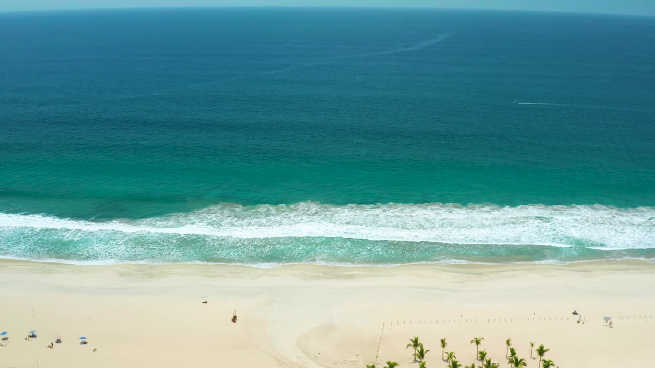 San Jose del Cabo, Mexico beach aerial with deep blue-green water in the upper frame, white surf breaking in a horizontal line, open sand below, and no visible people.