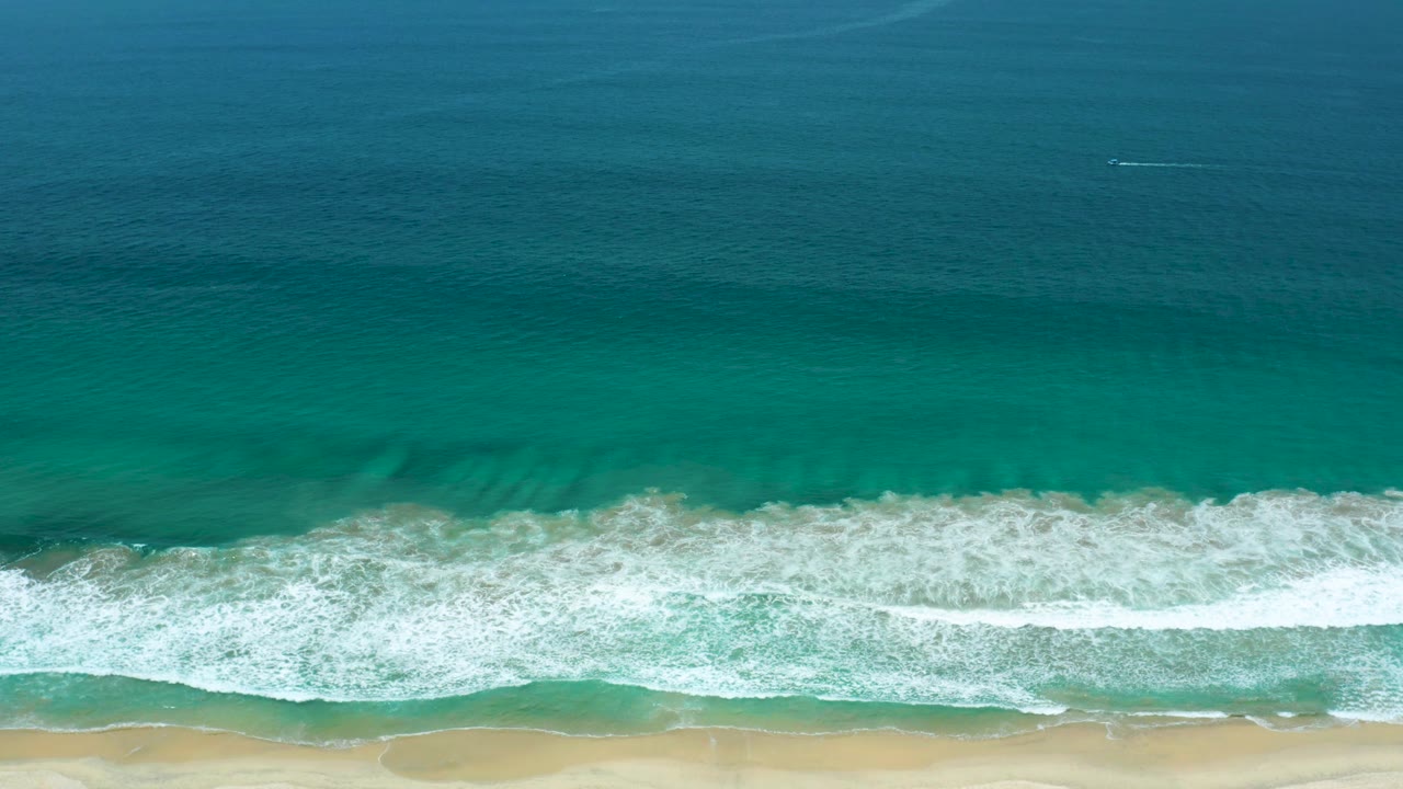 Top-down coastal frame with a central white surf band, darker teal water above, and pale sand below in San Jose Del Cabo, Mexico, with no visible people.