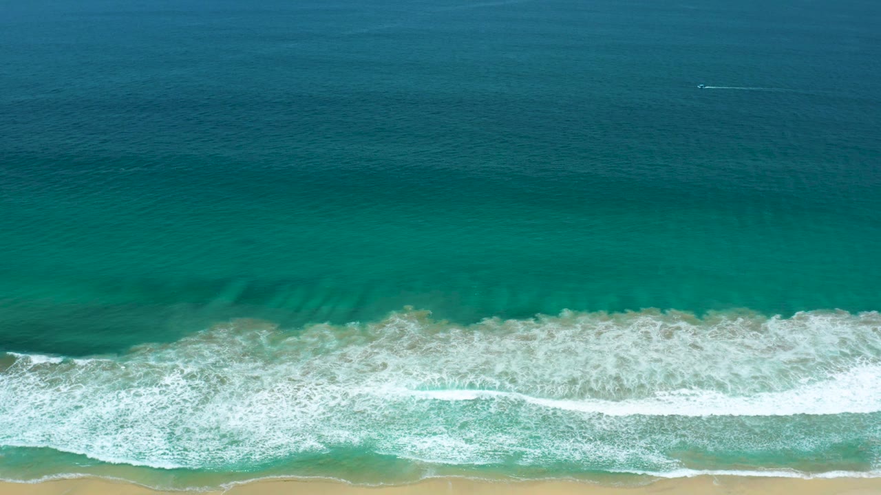 Teal ocean surface fills most of the frame above a breaking white wave line and a narrow sand strip in San Jose Del Cabo, Mexico, with no visible people.