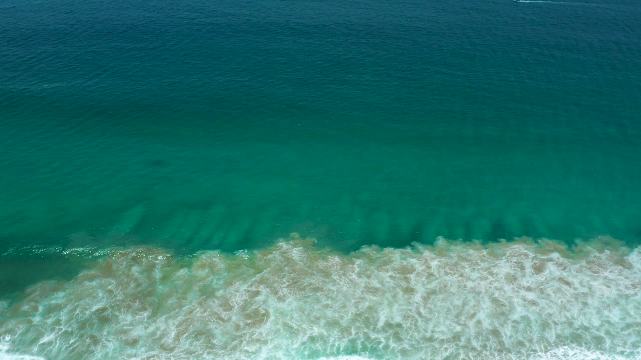 Top-down view of blue-green water and white surf reaching tan sand in San Jose Del Cabo, Mexico, with no visible people.