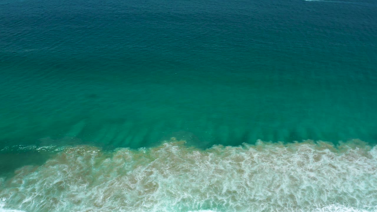 Aerial frame of green-blue sea with a broad white wave band rolling toward sand in San Jose Del Cabo, Mexico, with no visible people.