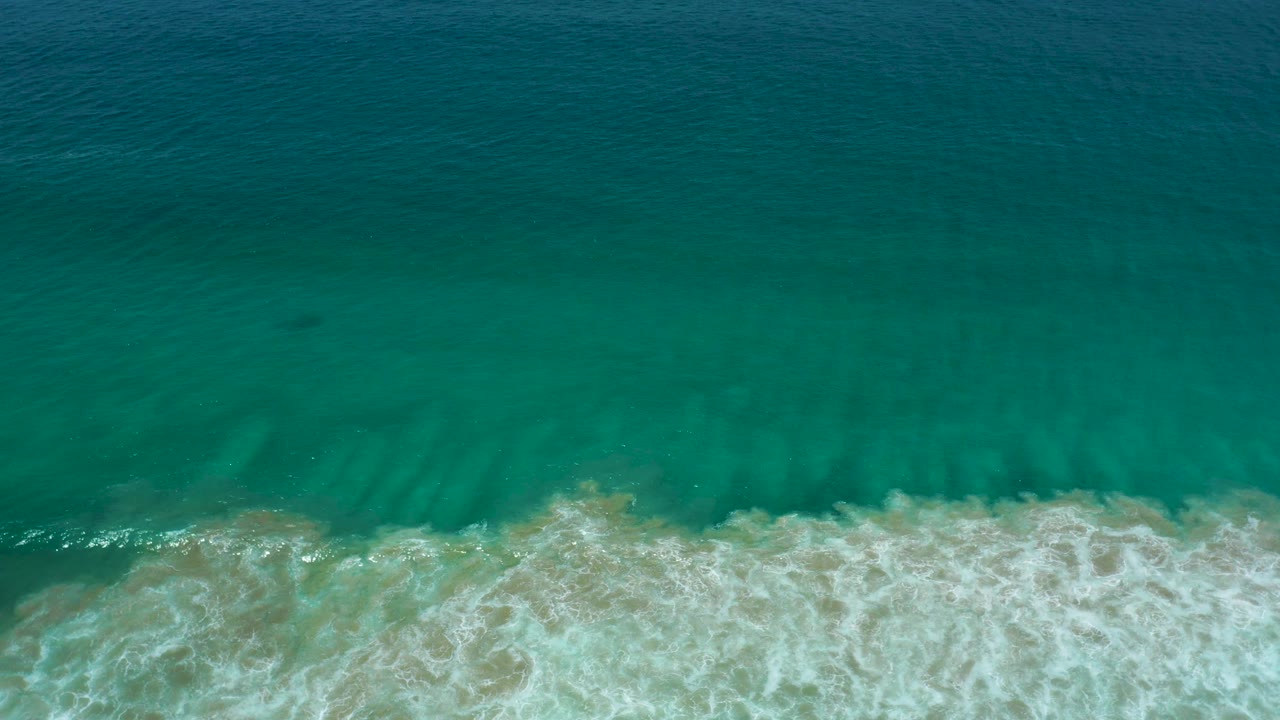 Overhead view of teal water above a foamy breaking wave near the sandy edge in San Jose Del Cabo, Mexico, with no visible people.