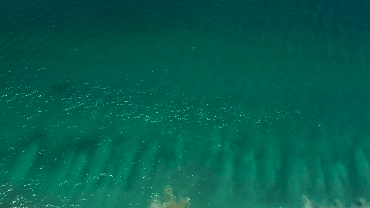 Aerial top-down frame of San Jose del Cabo seawater with green-blue color, soft surface ripples, pale underwater sand marks, and no visible people.