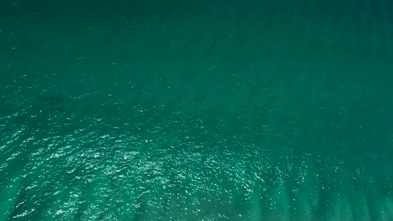 Aerial frame over dark teal San Jose del Cabo water with a broad patch of sun glare low in the image and no visible people.