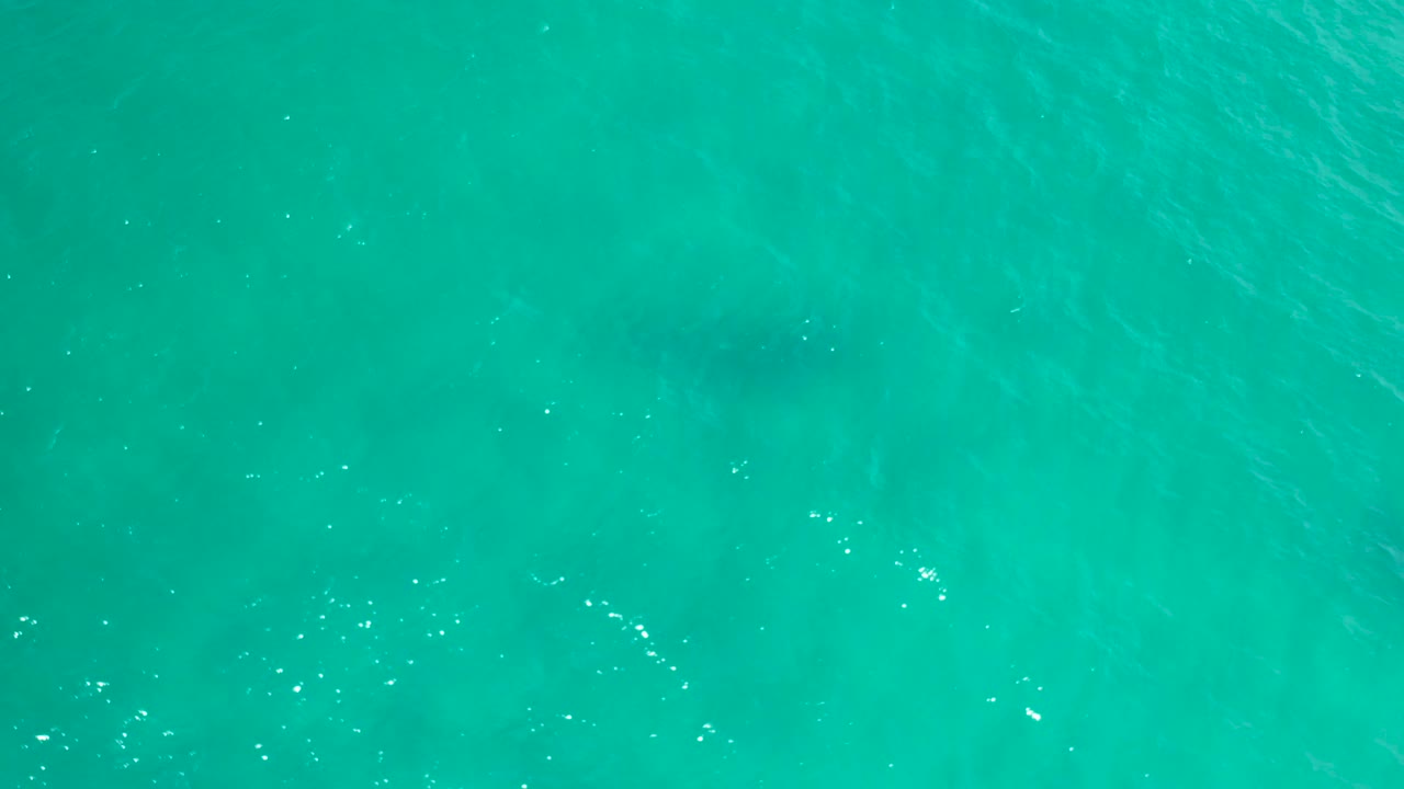 Aerial downward view of bright turquoise water with a soft dark underwater shape near the lower area and no visible people.