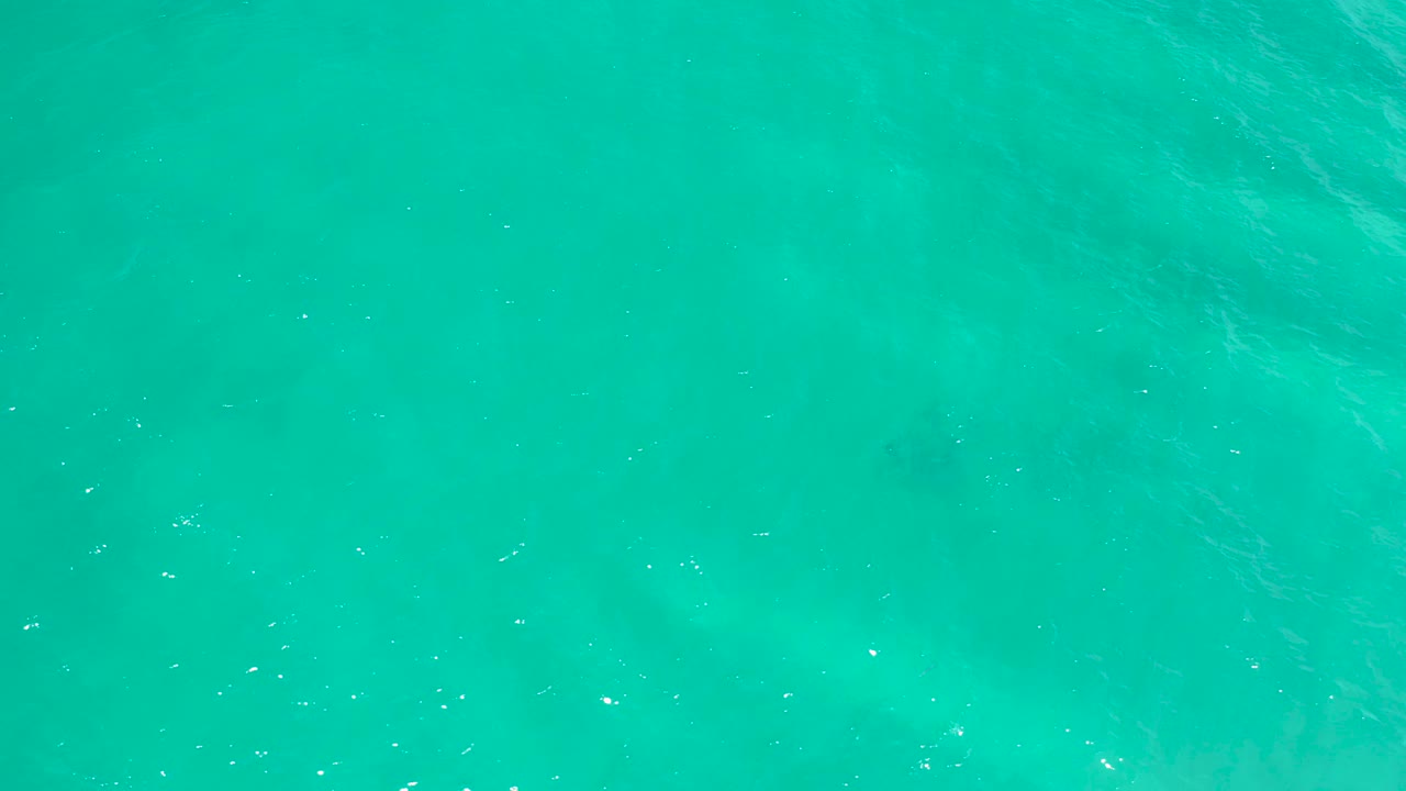 Bright aqua water in San Jose Del Cabo, Mexico shows scattered white surface flecks and faint submerged color shifts, with no visible people.