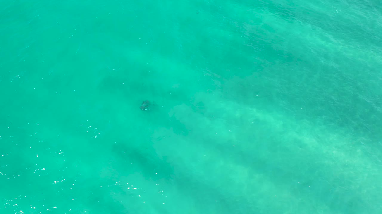 Turquoise San Jose Del Cabo water appears from above with pale shallows crossing the lower frame, faint dark shapes under the surface, and no visible people.